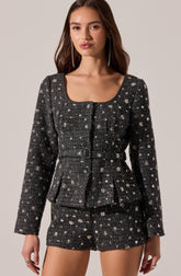A woman is wearing a matching two-piece outfit consisting of a long-sleeved, fitted jacket and a pair of shorts, both made from a dark, textured fabric adorned with a pattern of small, light-colored floral or star-like motifs. The jacket has a round neckline, button closures down the front, and a belt at the waist. The shorts are tailored with a clean, simple design.